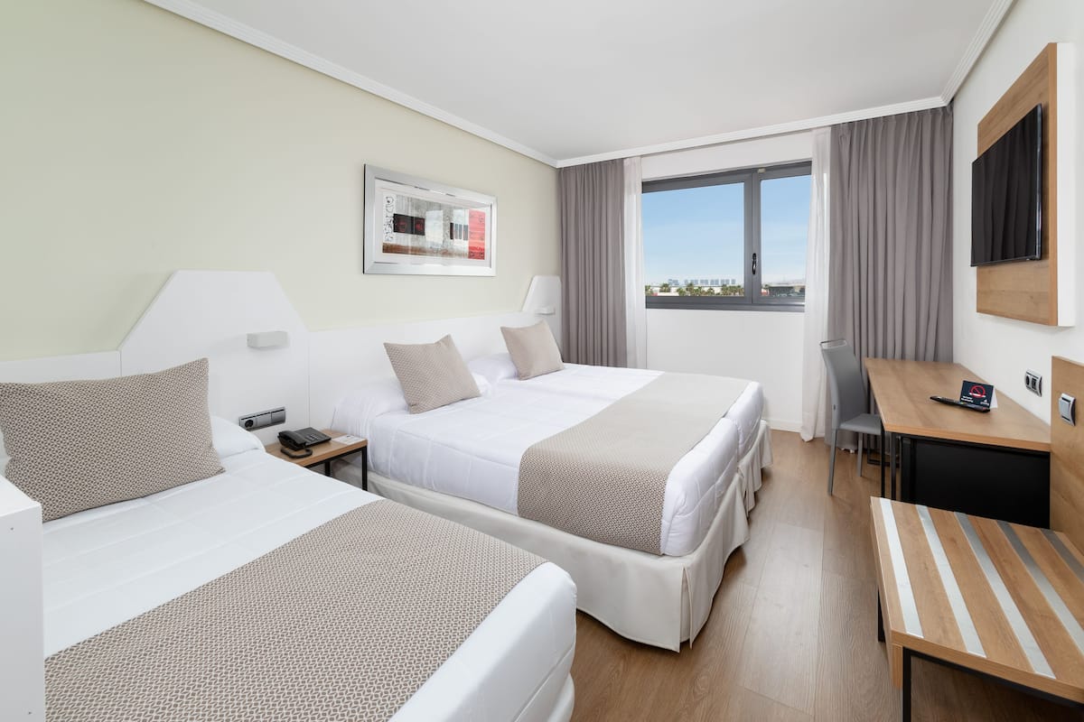 Ein Hotelzimmer mit zwei Betten, einem Schreibtisch, einem Stuhl, einem Fernseher und einem Fenster mit Blick auf die Stadt.