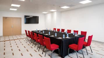 Un salón de conferencias con una larga mesa, sillas rojas, botellas de agua y un televisor de pantalla plana en la pared.