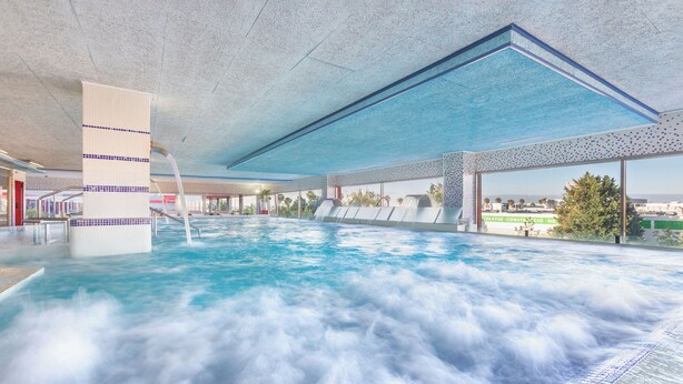 Une piscine intérieure agrémentée d’une cascade, avec une eau d’un bleu cristallin et une vue sur le paysage urbain grâce à de grandes fenêtres.
