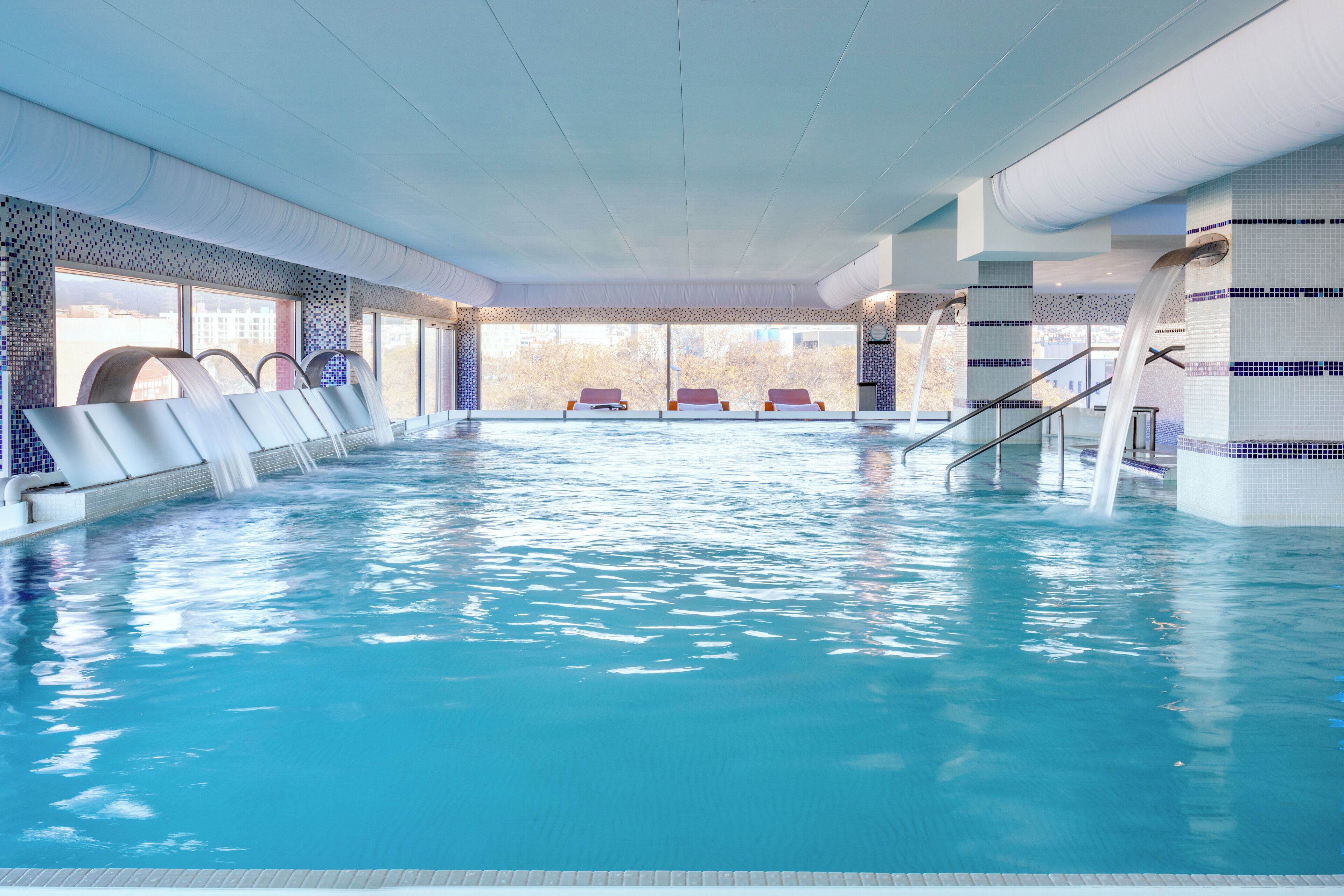 An indoor swimming pool with clear water, lounge chairs, and a view of the outside.