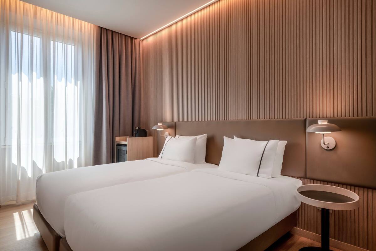 A hotel room with a large bed, two bedside tables, a nightstand, and a wall-mounted lamp.