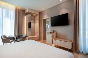 A modern hotel room with a large bed, a desk with a chair, a TV mounted on the wall, and a vanity with a mirror.