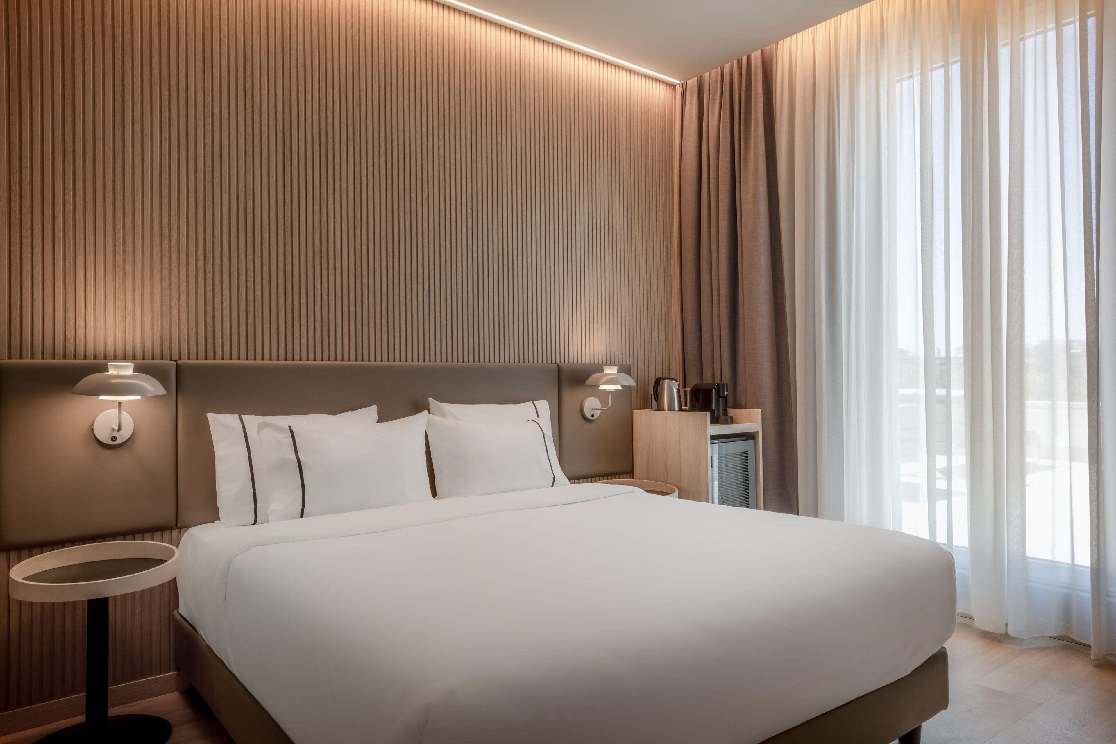A modern hotel room with a large bed, two bedside tables, a nightstand, and a view of the outdoors through sheer curtains.