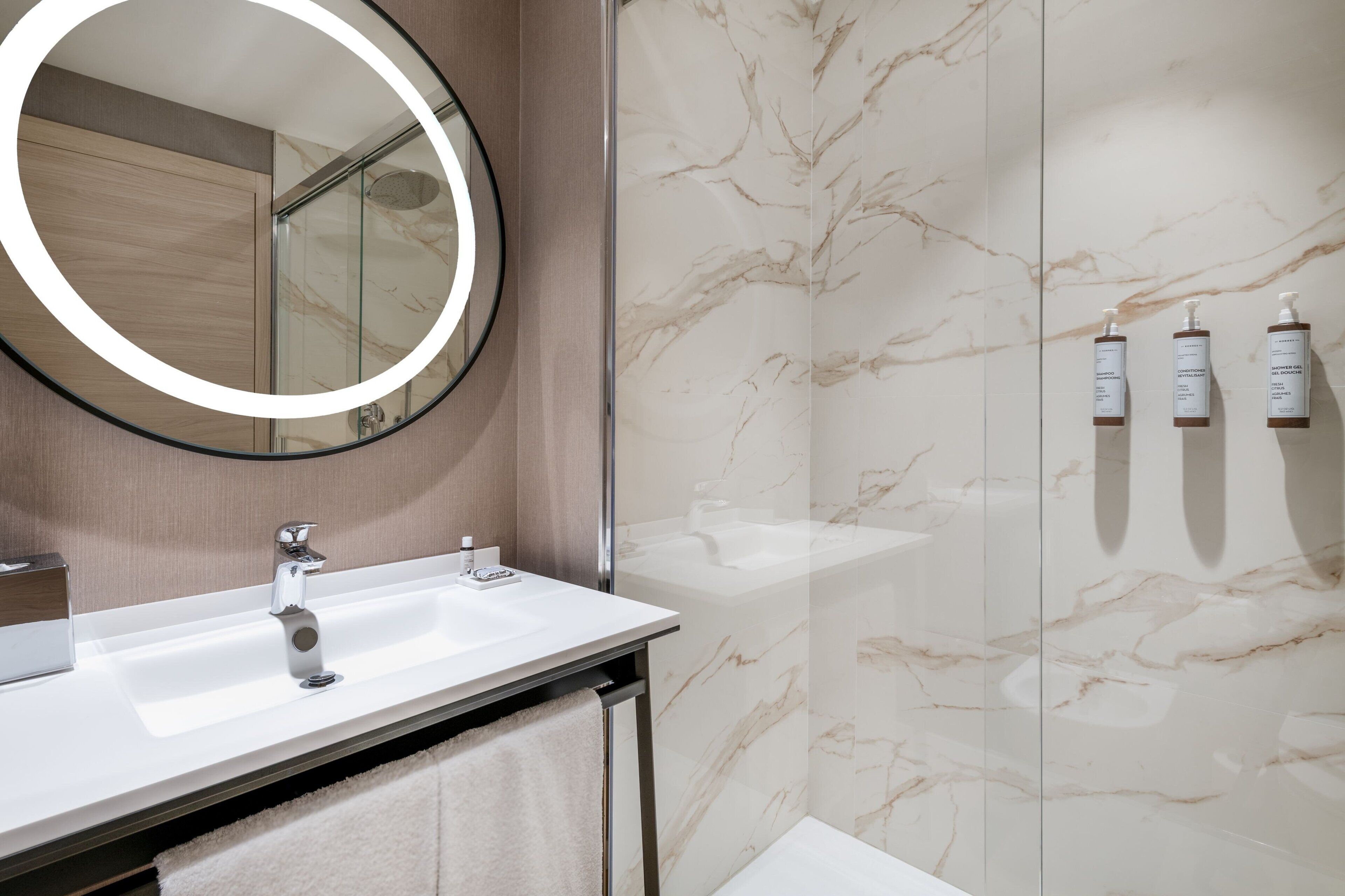 Un baño moderno con un espejo redondo, un lavamanos blanco y una cabina de ducha de vidrio.