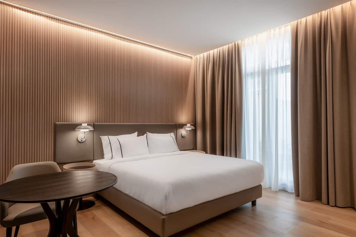 A modern hotel room with a large bed, a bedside table, a chair, and a lamp.