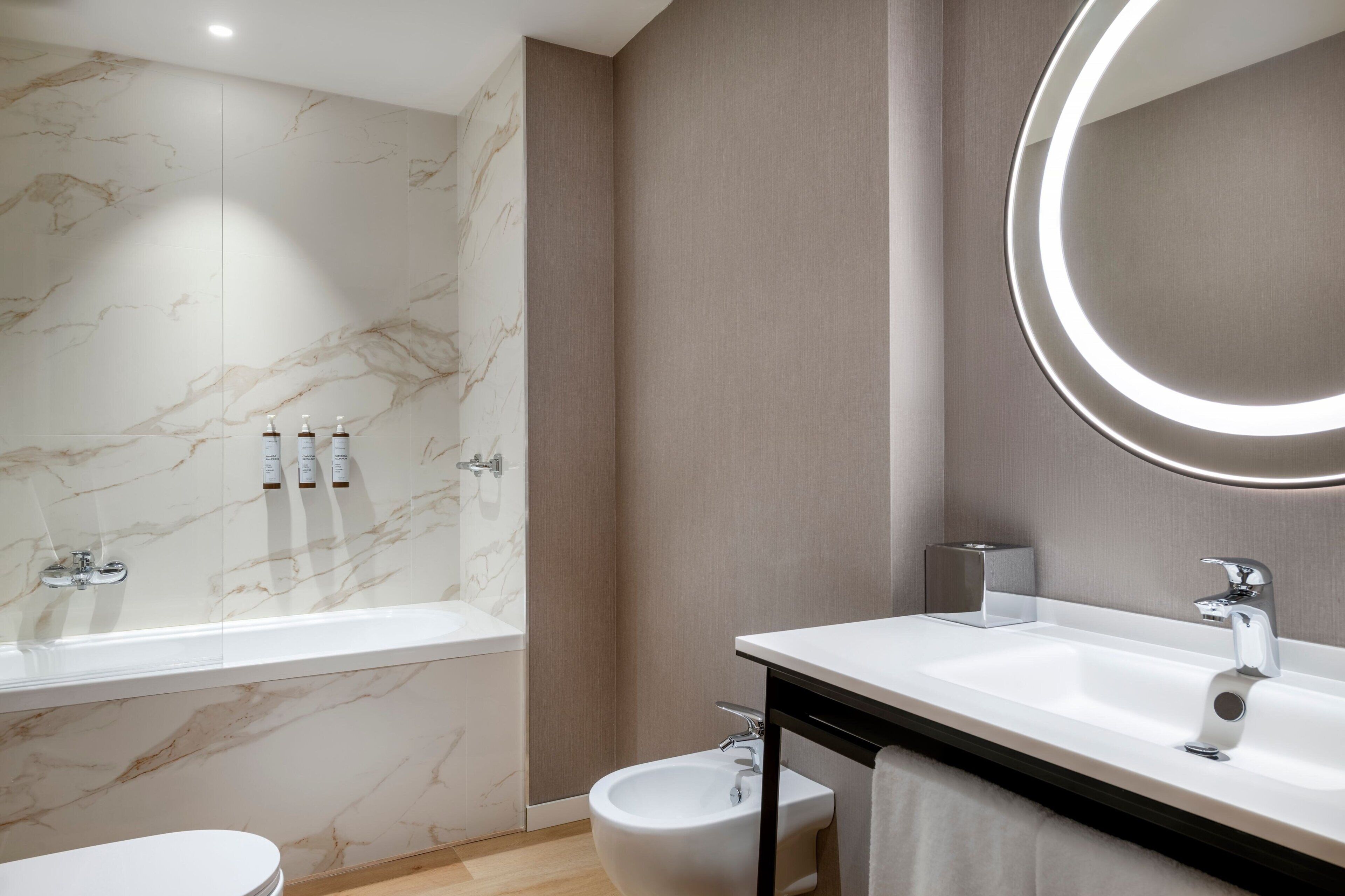 Une salle de bain moderne avec un mur en marbre, une baignoire sur pieds, un bidet et un grand miroir doté d’un éclairage circulaire.