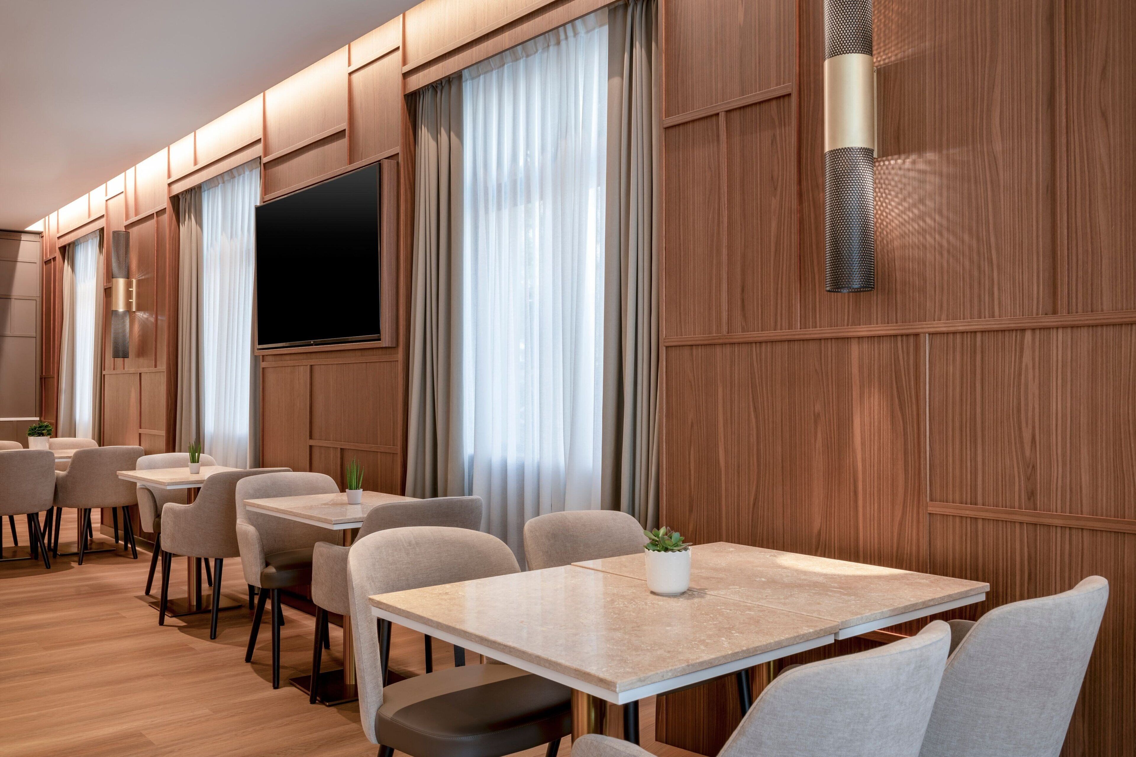 A modern hotel room with wooden paneling, a flat-screen TV, a large window with curtains, and a seating area with tables and chairs.