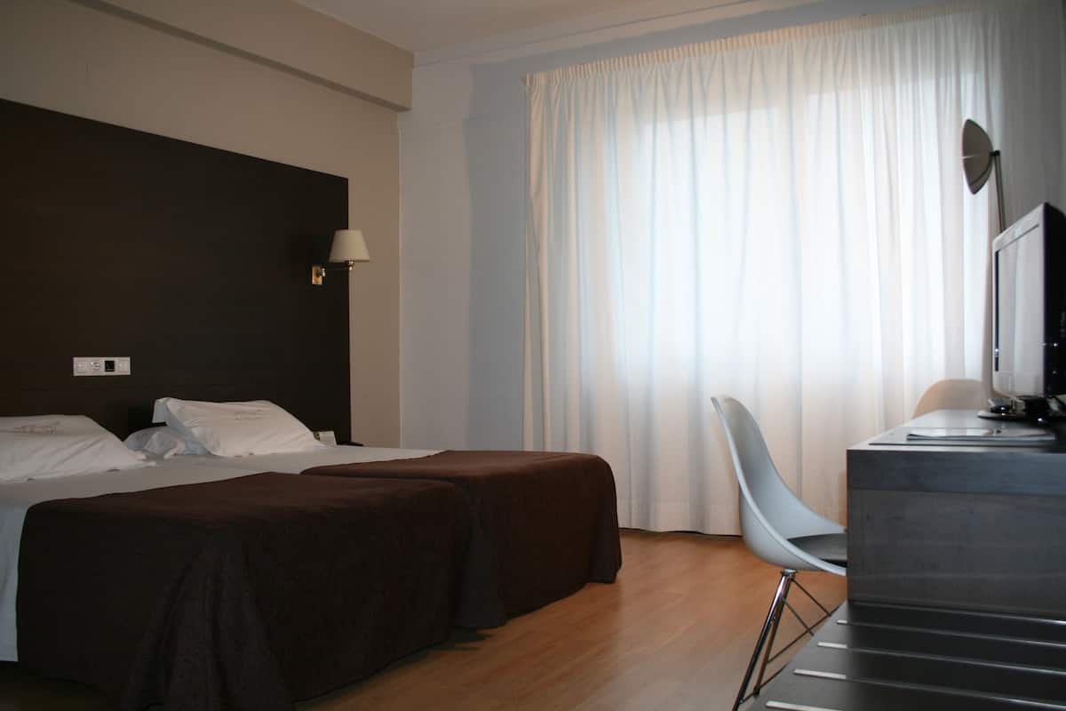 A hotel room with a bed, two bedside tables, a desk with a chair, a TV, and a window with curtains.