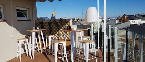 A balcony with white bar stools and a table, overlooking a cityscape with buildings and a clear sky.