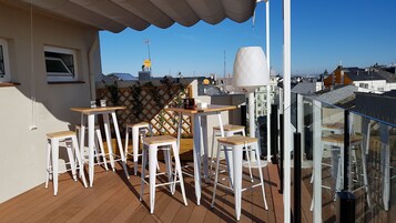 Un balcón con taburetes blancos y una mesa, con vistas a un paisaje urbano de edificios y cielo despejado.