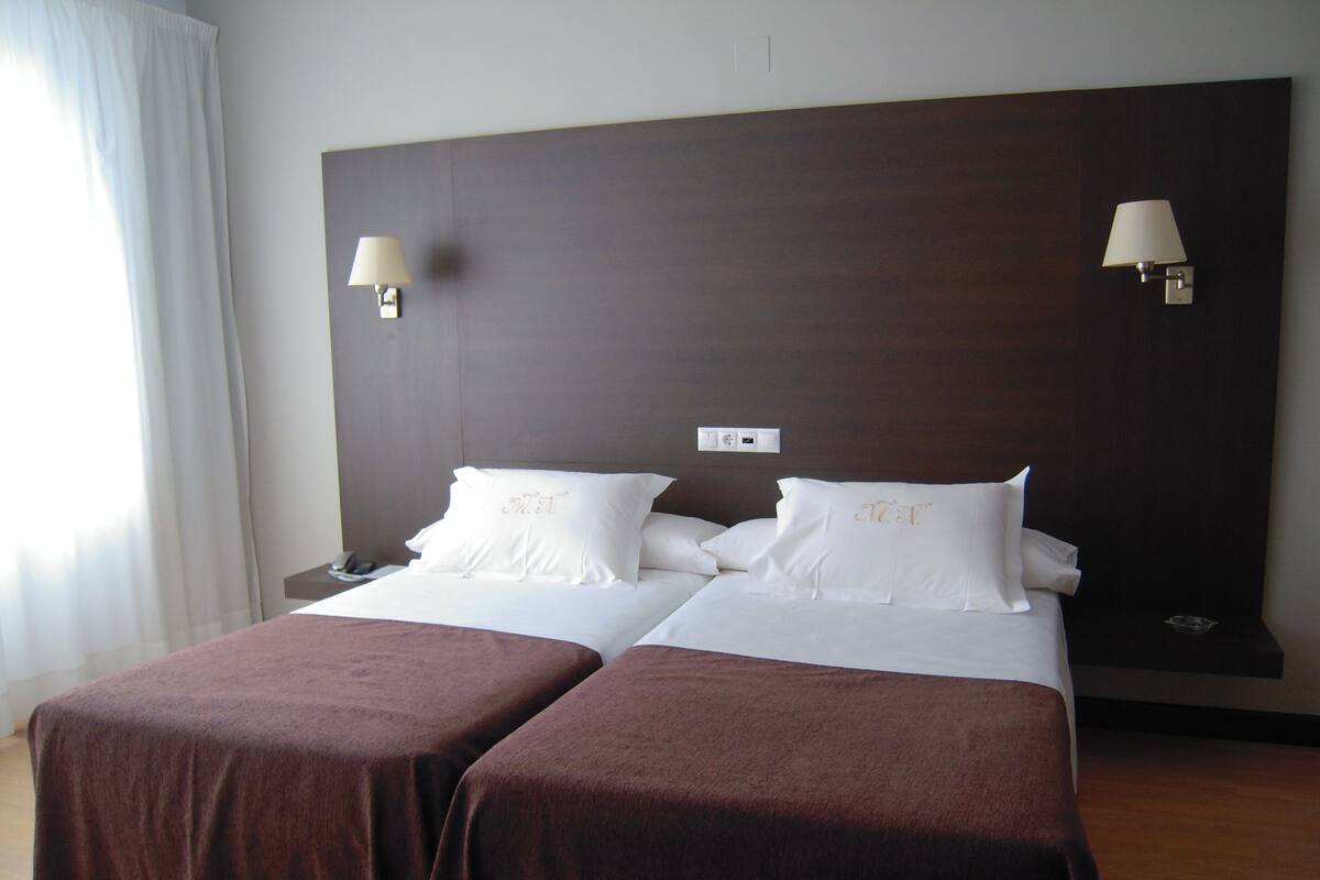 A hotel room with two beds, each with a brown bedspread and white linens, two wall-mounted lamps, and a nightstand with a phone.