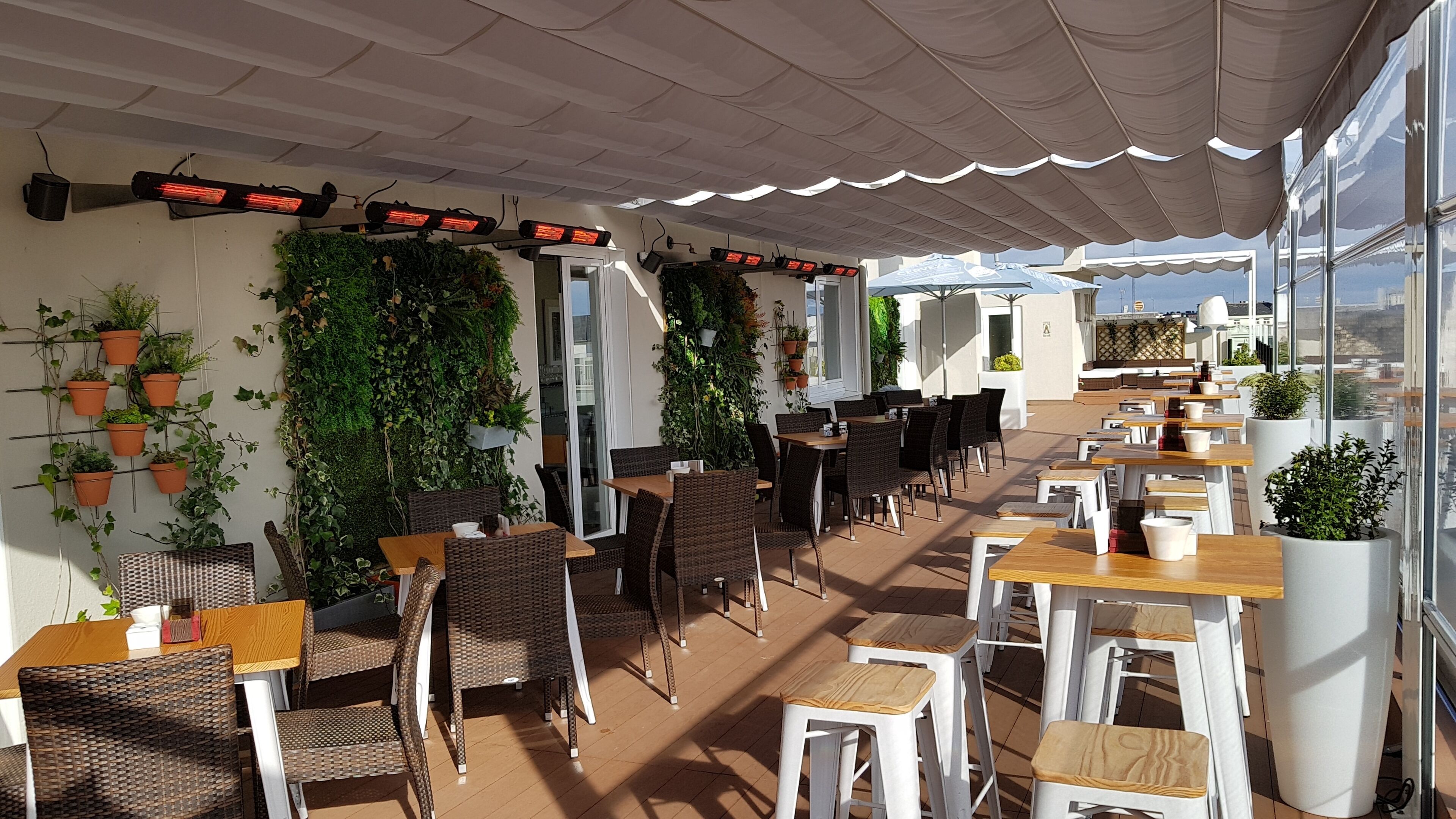 Een terras met rieten stoelen en tafels, bloempotten en een plafond met verlichting.