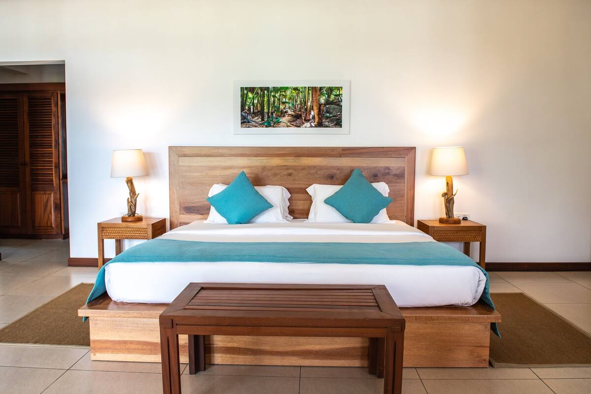 A neatly made bed with a wooden headboard, two nightstands with lamps, a bench, and a framed picture on the wall.