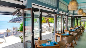 A seaside restaurant with glass doors leading to a beach view, wooden tables, and wicker chairs. - Hotel L'Archipel (Praslin Island)