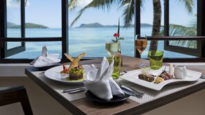 A table set with plates of food, glasses of wine, and a view of the ocean through large windows. - Hotel L'Archipel (Praslin Island)
