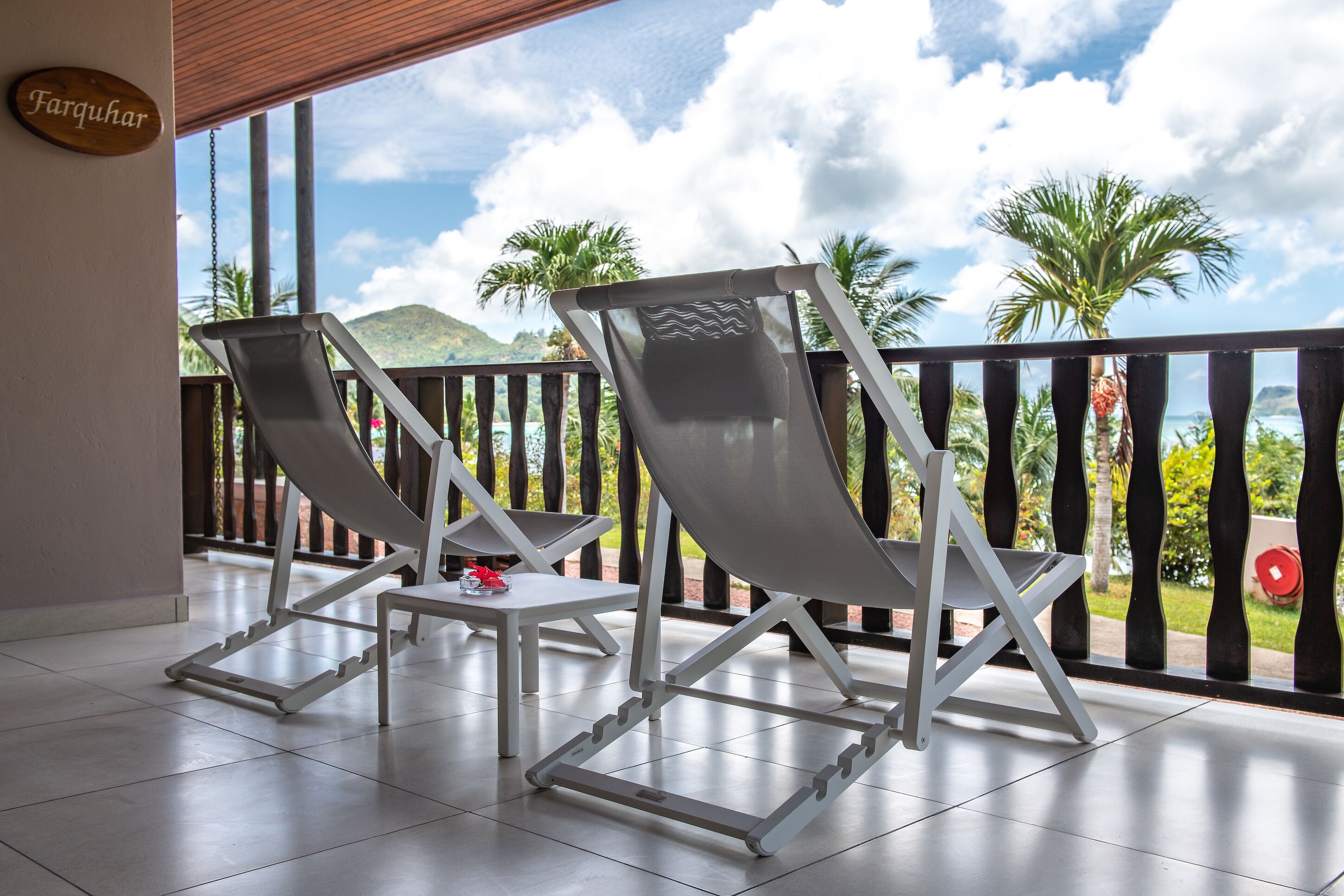 A balcony with two white lounge chairs and a small table, overlooking a scenic view with palm trees and mountains.