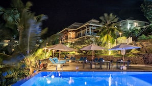 A swimming pool with lounge chairs and umbrellas, surrounded by palm trees and a stone wall. - Hotel L'Archipel (Praslin Island)