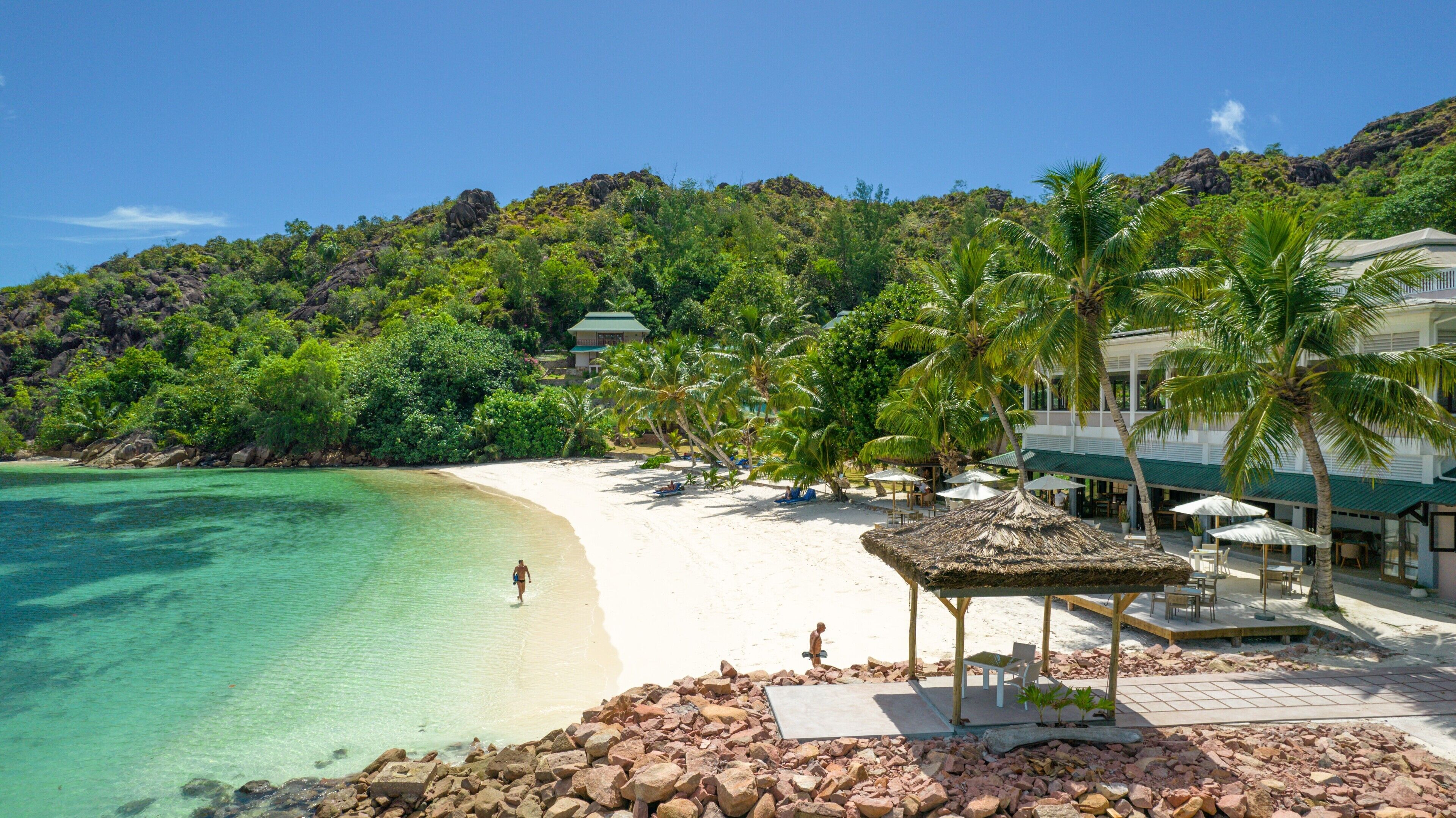 Ein Strandresort mit Sandstrand, klarem türkisblauem Wasser und üppiger Vegetation. Es gibt Palmen, ein strohgedecktes Pavillon und ein Gebäude mit Gründach.