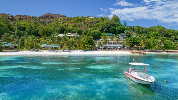 A tropical beach resort with clear turquoise waters, a boat docked near the shore, and a backdrop of lush greenery and a mountain. - Hotel L'Archipel (Praslin Island)