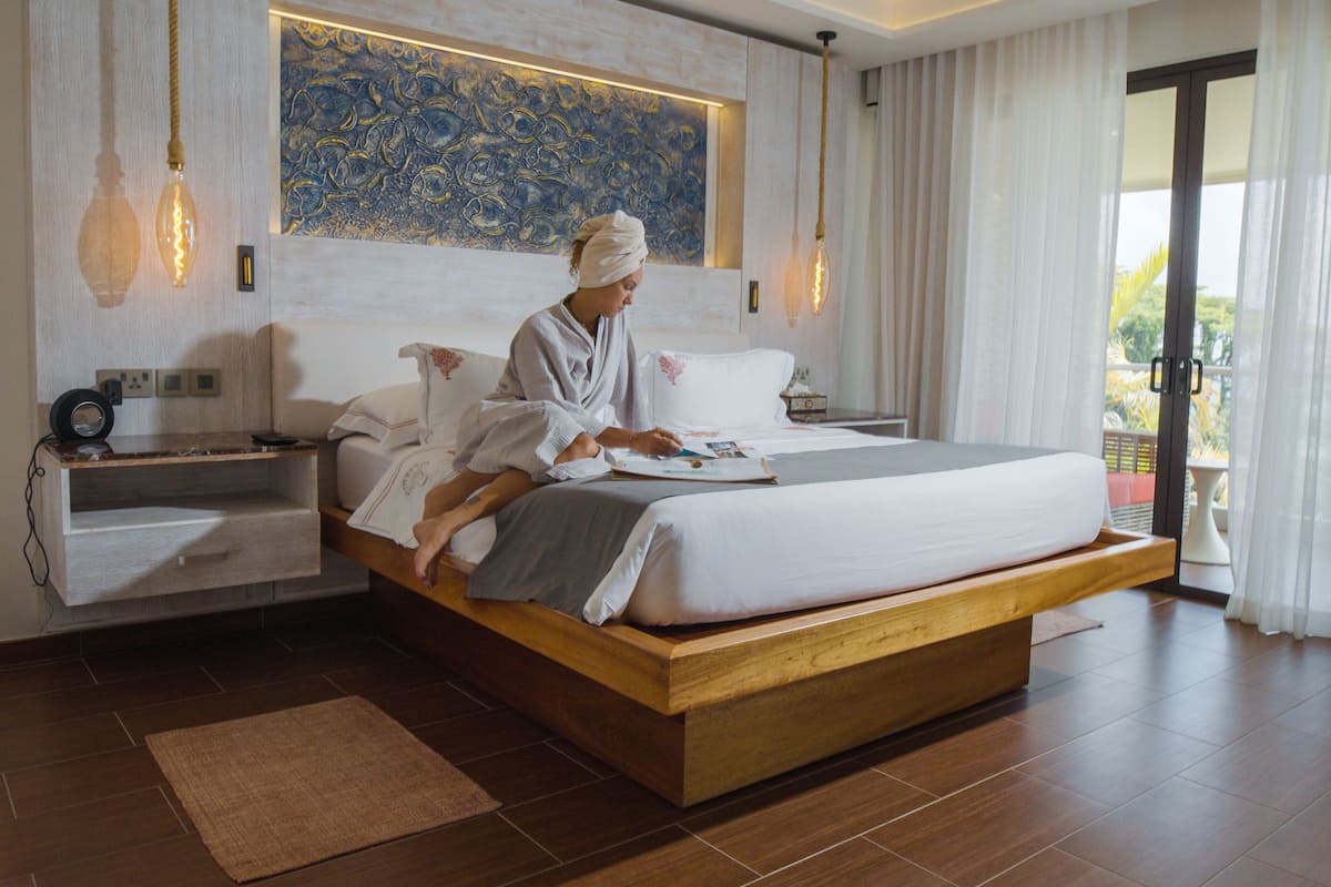 Ein modernes Schlafzimmer mit einem großen Bett, einem Nachttisch, einer Wandleuchte und Blick ins Freie durch eine Glastür.