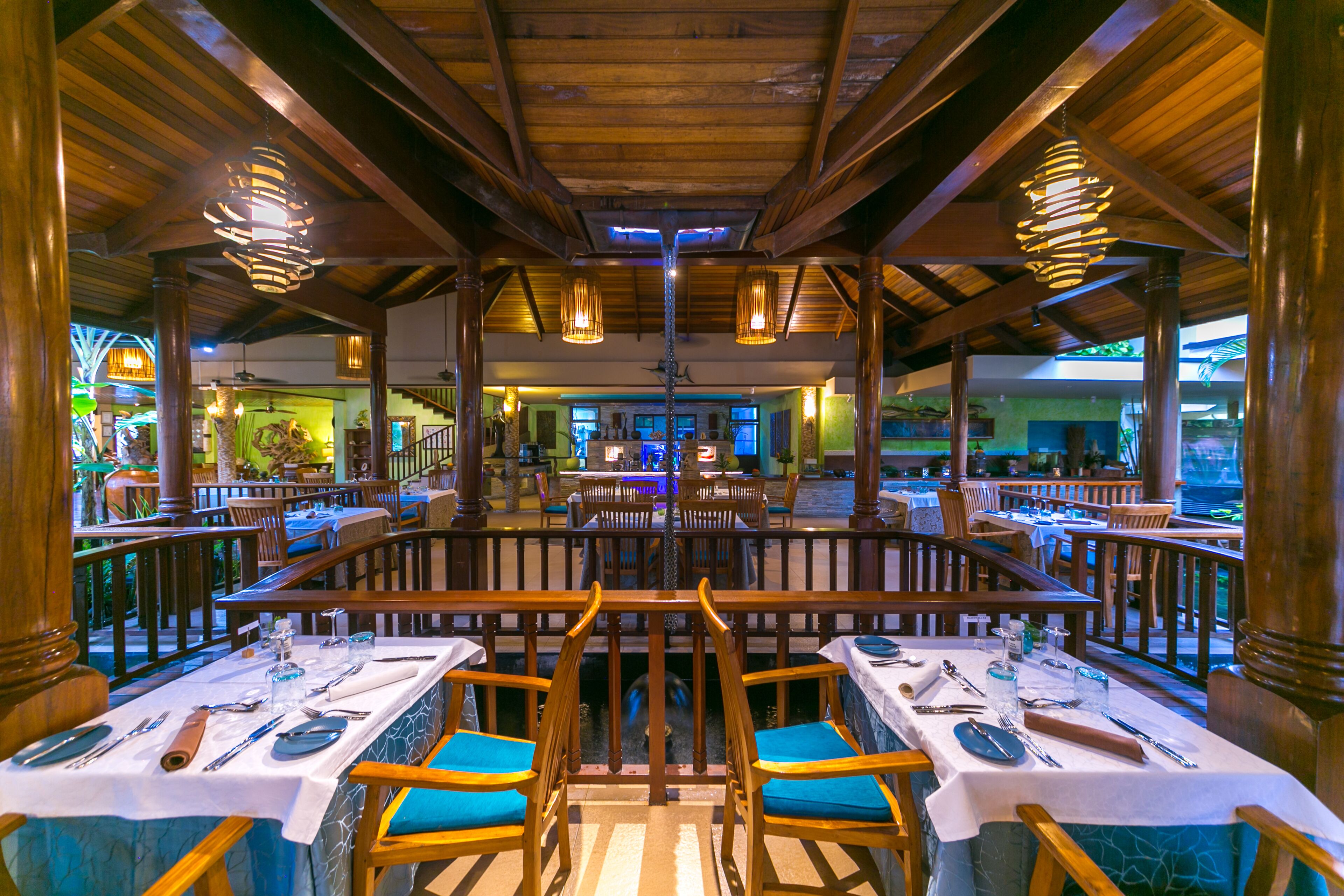 A restaurant with wooden beams and hanging lights, tables set with cutlery, and blue cushioned chairs.