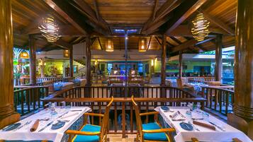 A restaurant with wooden beams and hanging lights, tables set with cutlery, and blue cushioned chairs.