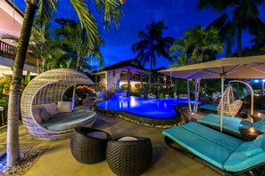 Area piscina con poltrone prendisole, una sedia a dondolo e una piscina ben illuminata, circondata da palme.
