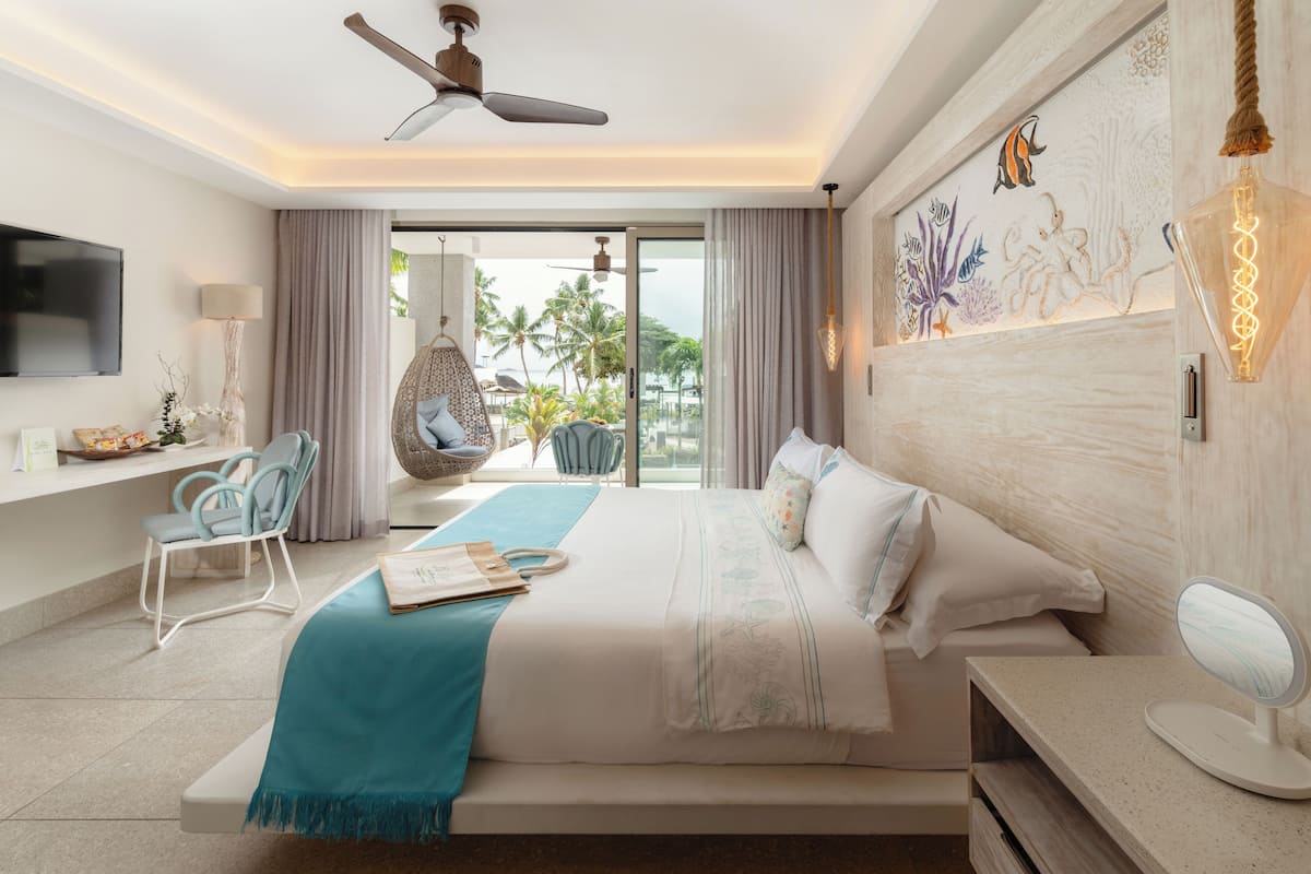Ein modernes Hotelzimmer mit einem großen Bett, einem Deckenventilator, einem Fernseher, einer Sitzecke mit einem Sessel und einem kleinen Tisch sowie Blick auf Palmen durch das Fenster.