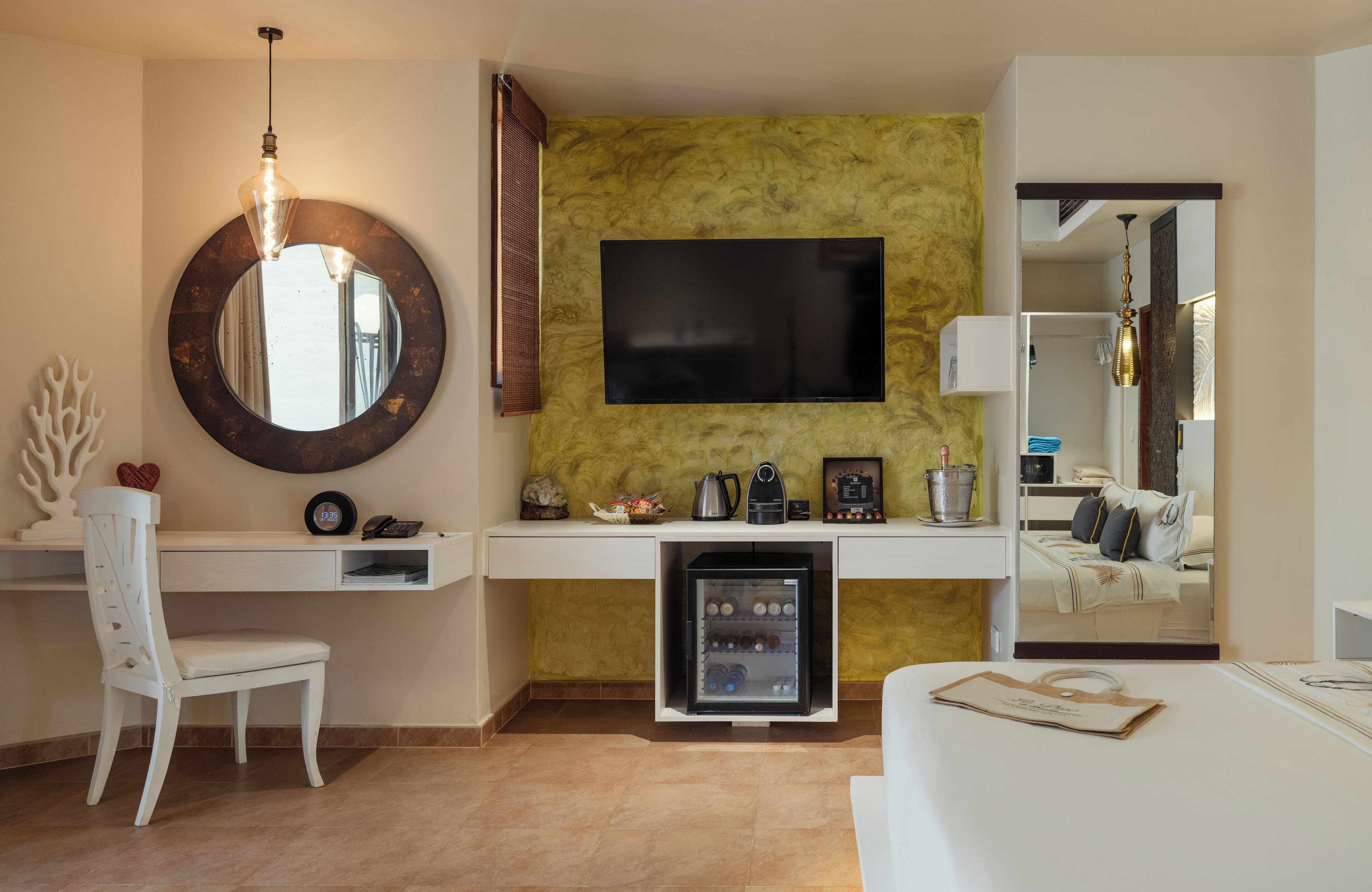 A hotel room with a round mirror, a flat-screen TV, a minibar, a white desk, a white chair, and a bed.