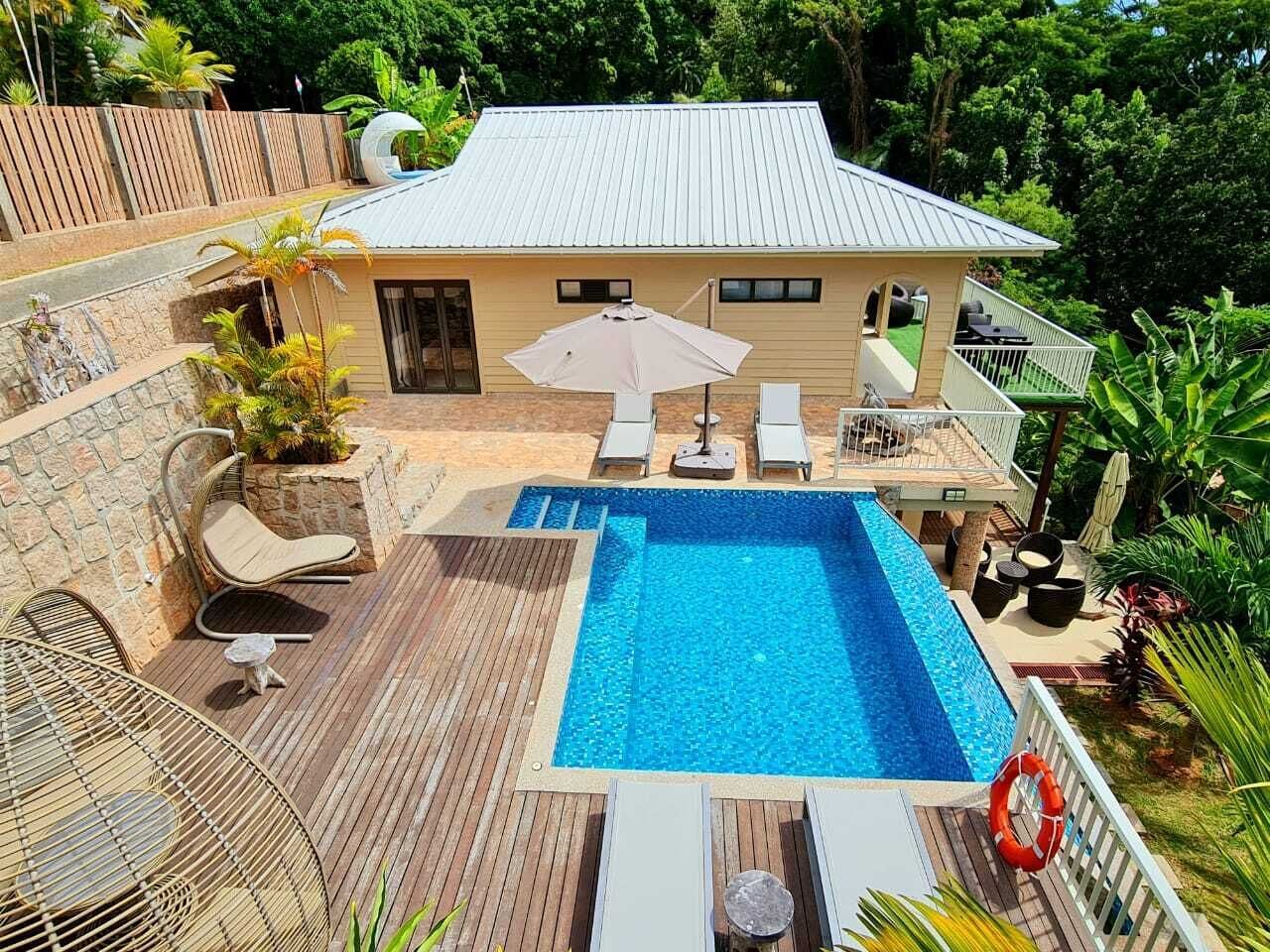 Una piscina con lettini e un ombrellone, circondata da un pontile in legno e una casa con tetto in metallo.