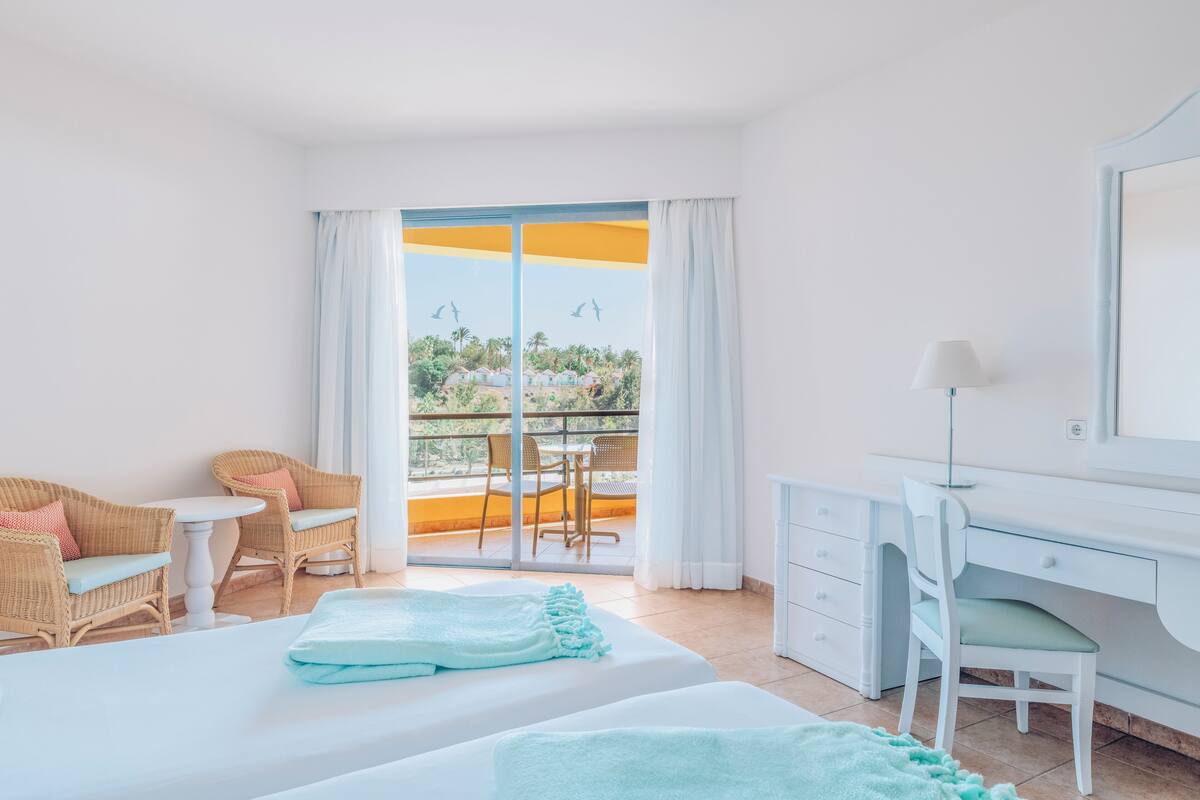 A hotel room with a balcony, a desk, two wicker chairs, a bed, and a small table.