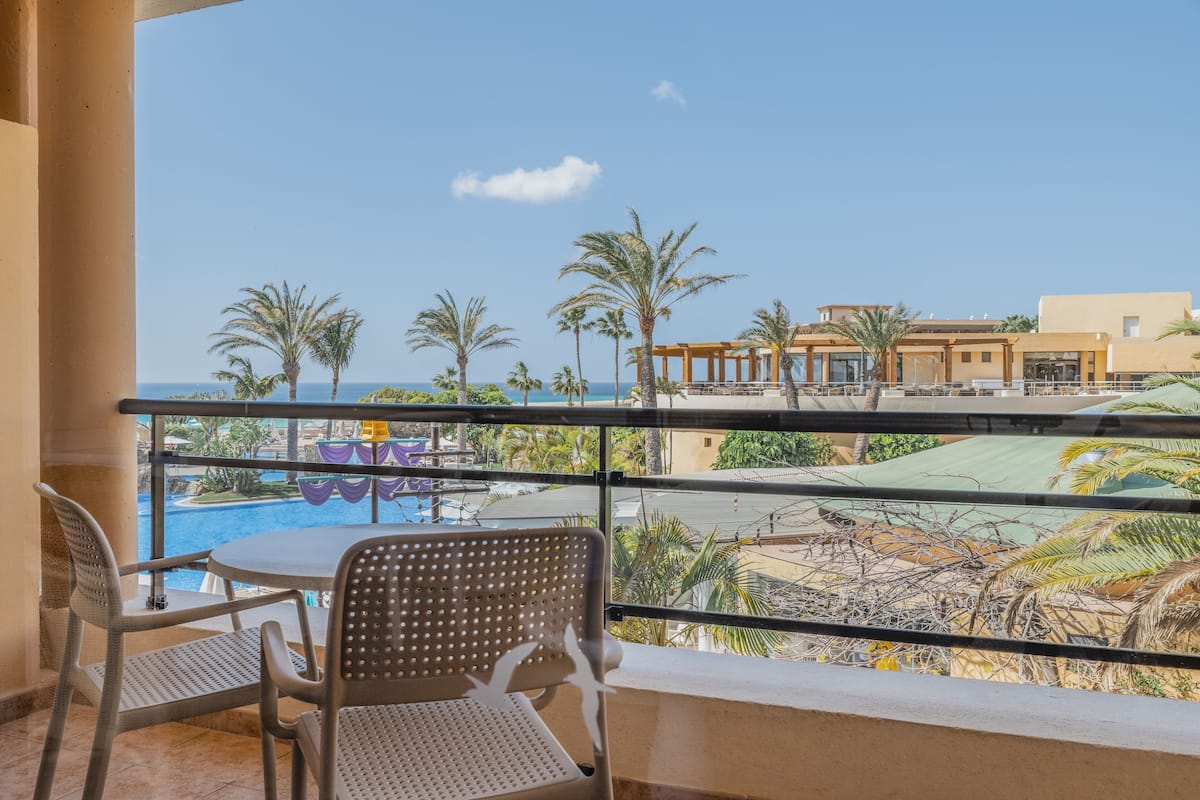 A balcony with a view of a pool and palm trees, a table with chairs, and a building in the background.