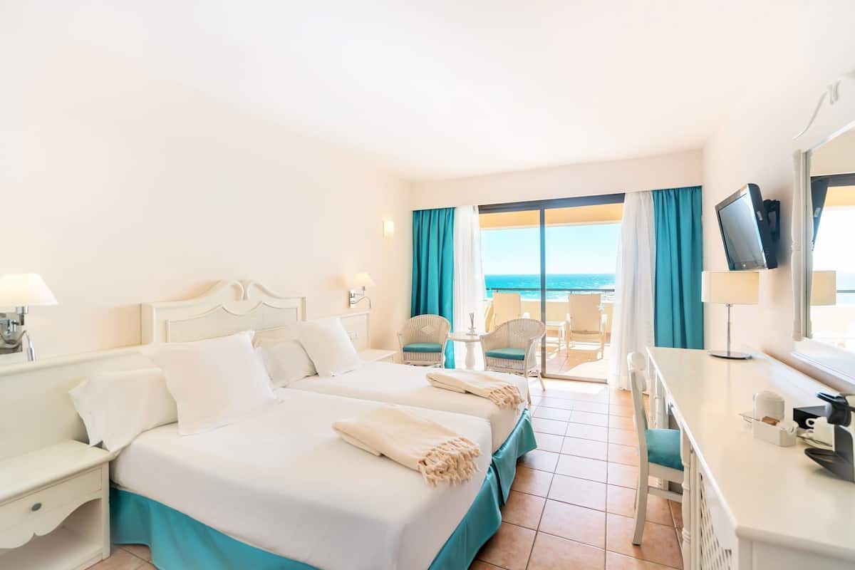 A hotel room with two beds, a balcony with a view of the sea, a desk with a TV, and a chair.