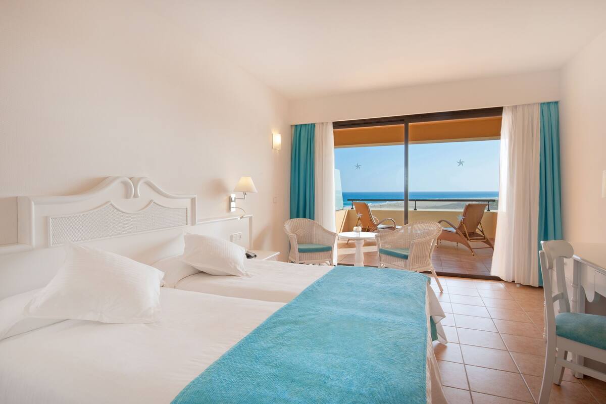A hotel room with two beds, a balcony with a view of the sea, and a table with chairs.