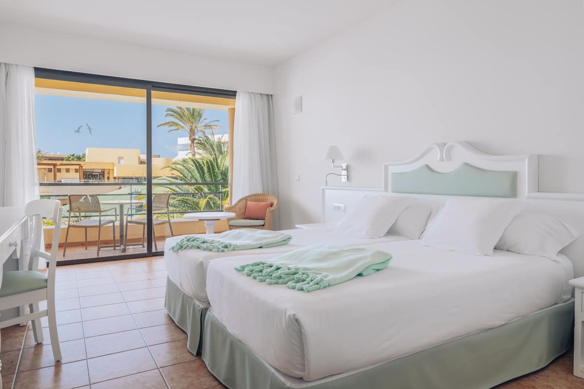 A hotel room with two beds, a balcony with a table and chairs, and a view of palm trees and buildings.