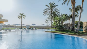 Ein Swimmingpool mit Liegestühlen und Palmen, umgeben von Gebäuden und blauem Himmel.