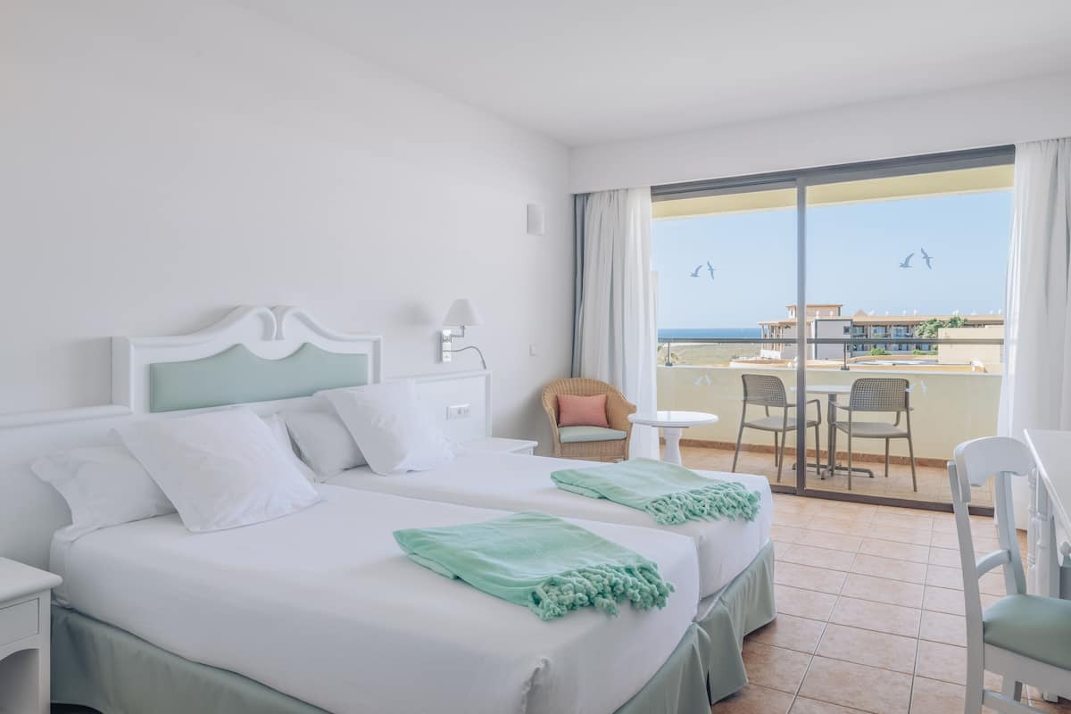 A hotel room with two beds, a balcony with a view of the sea, and a dining area with chairs and a table.