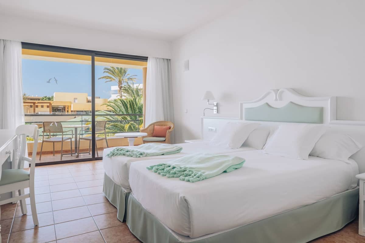 A hotel room with a balcony, two beds, a dining table, and chairs.
