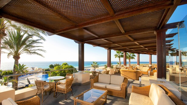 A covered outdoor seating area with wicker furniture, a glass table, and a view of the ocean. - Iberostar Waves Playa Gaviotas -All Inclusive (Pajara)