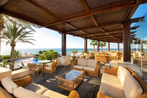 A covered outdoor seating area with wicker furniture, a glass table, and a view of the ocean. - Iberostar Waves Playa Gaviotas -All Inclusive (Pajara)