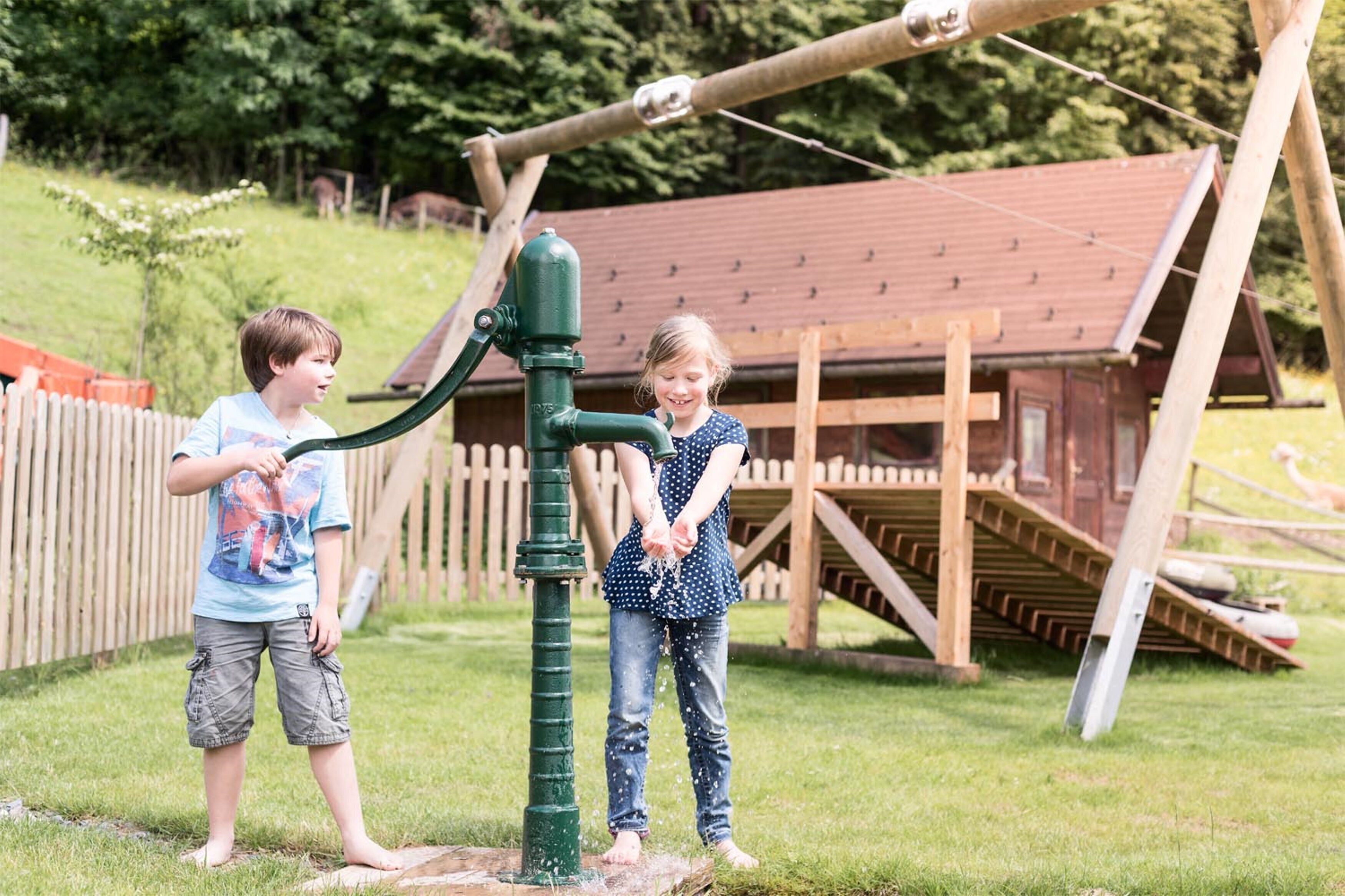 Two children playing with a water pump in a wooden play area with a wooden structure and a fenced garden in the background.