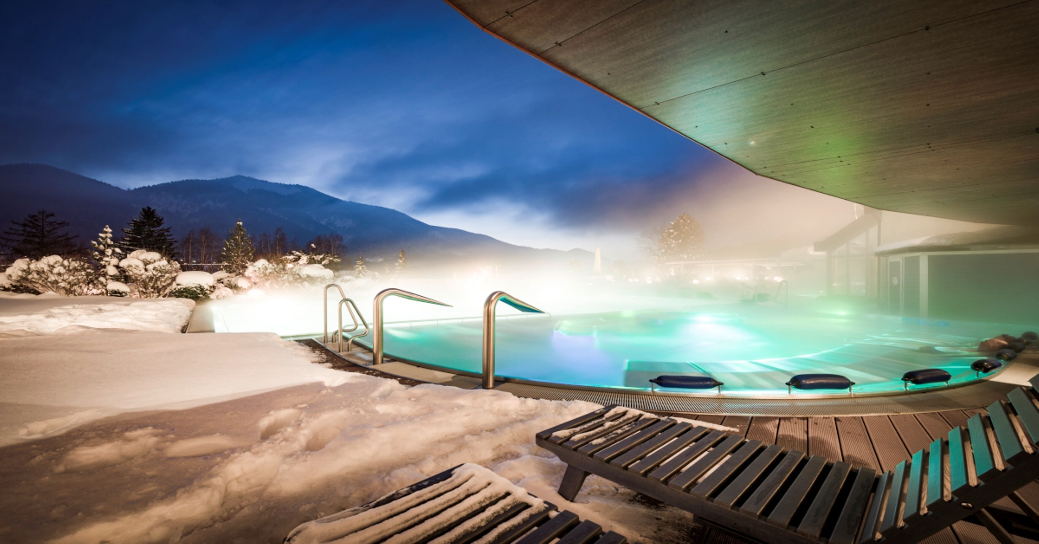 Vasca idromassaggio con vista su montagne innevate e terrazza in legno con poltrone relax.