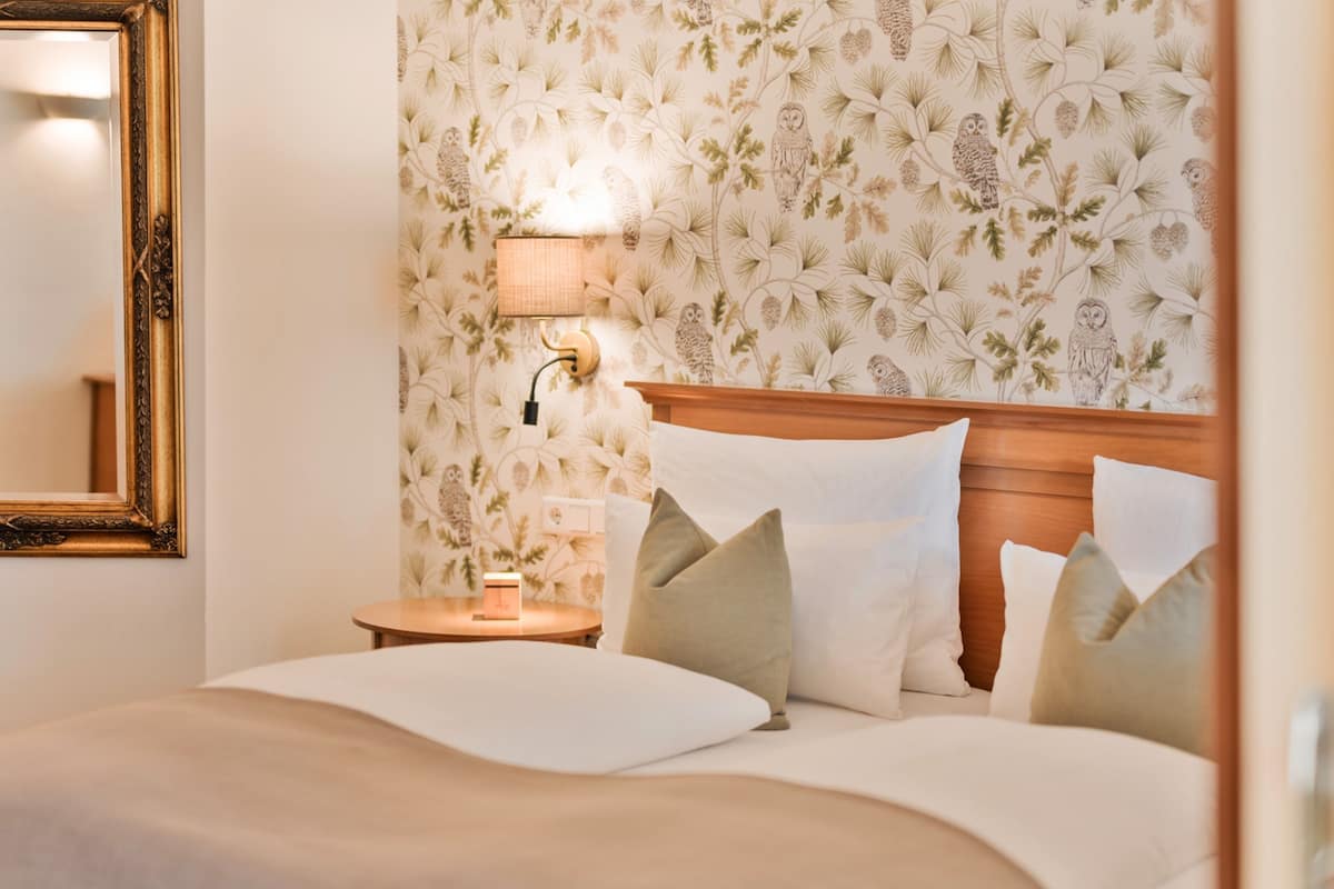 Un lit bien fait, avec du linge de lit blanc et une tête de lit en bois, une table de chevet avec une lampe et un papier peint à motifs floraux.