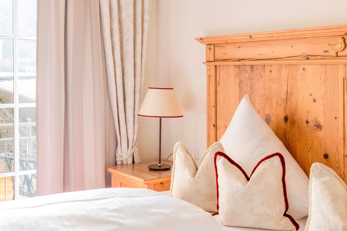 Un lit bien fait, avec du linge de lit blanc et des coussins décoratifs, une table de chevet en bois avec une lampe, et une fenêtre avec des rideaux.