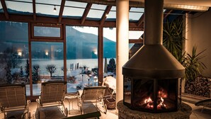 Un rincón exterior acogedor con chimenea de piedra, sillas de madera y vistas a un paisaje nevado a través de grandes ventanales.
