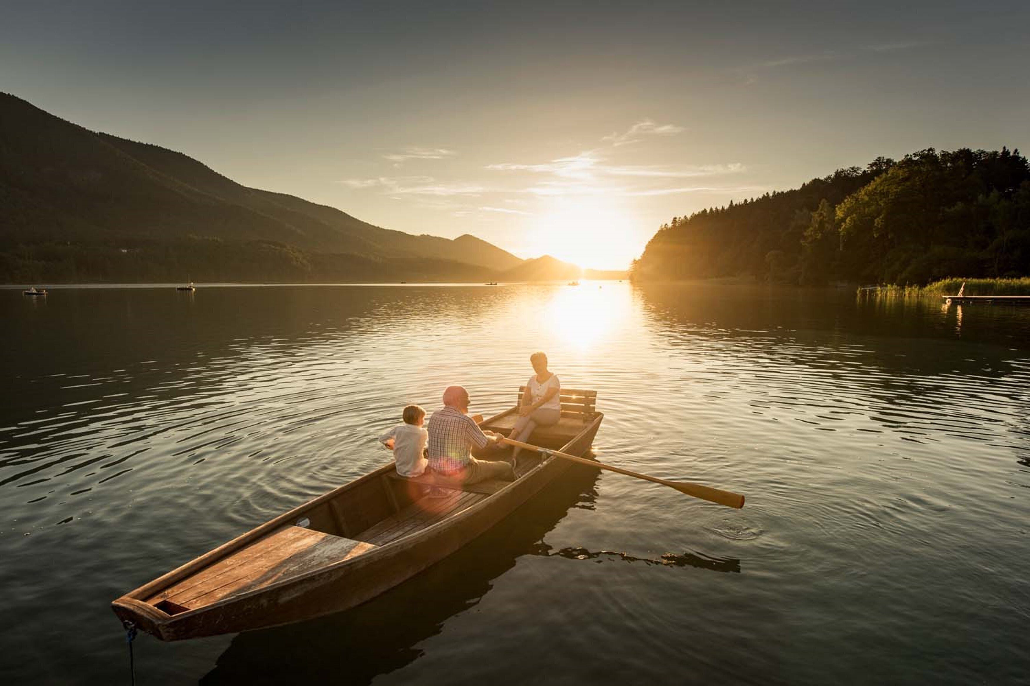 A wooden rowboat with three people on a calm lake at sunset.