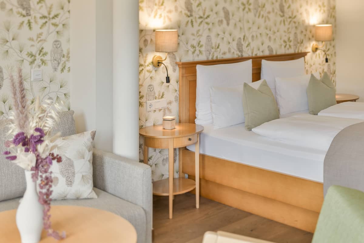Une chambre d’hôtel avec un lit, une table de chevet, une chaise et une petite table sur laquelle se trouve un vase de fleurs.