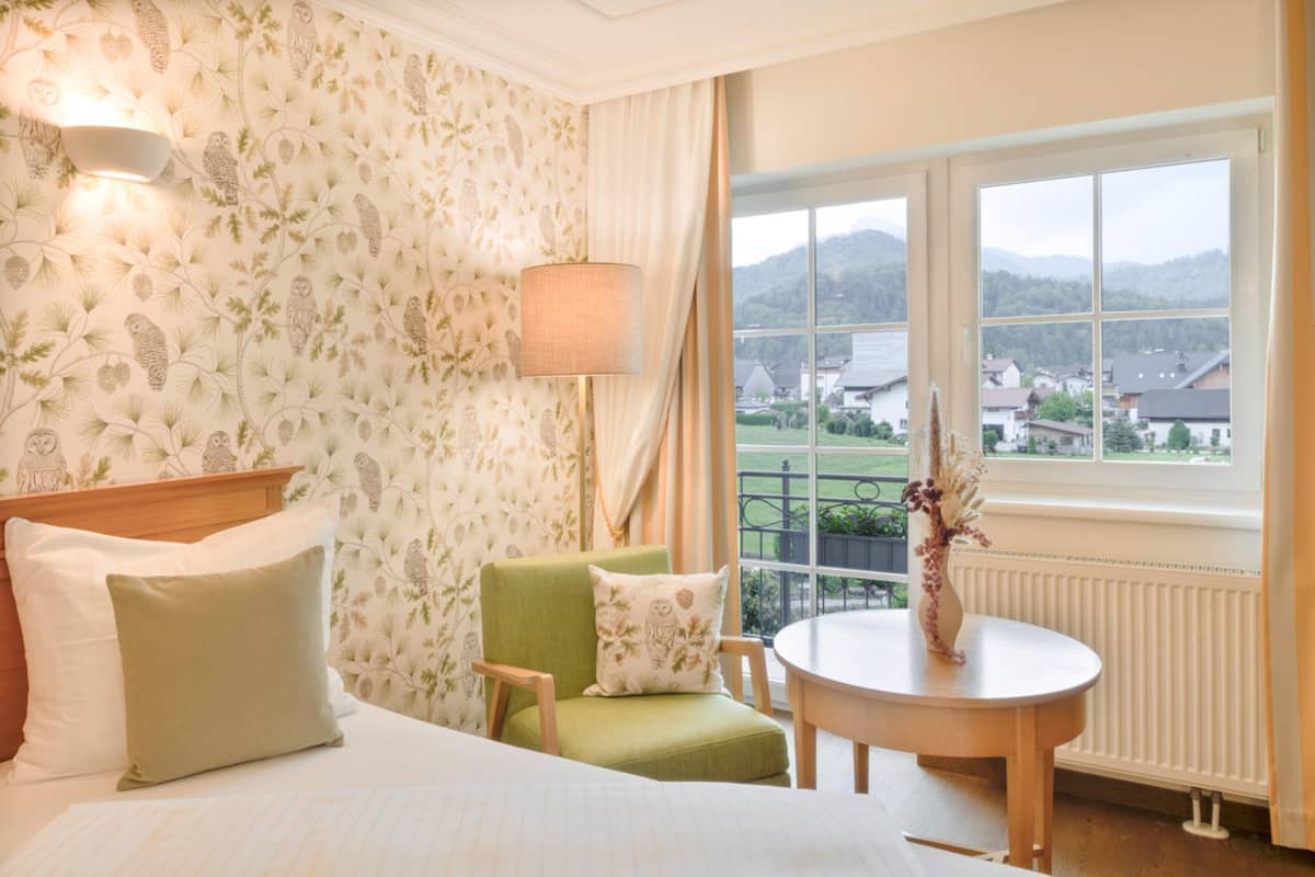 Une chambre d’hôtel avec un lit, un fauteuil vert, une table ronde et une vue sur les maisons par une grande fenêtre.