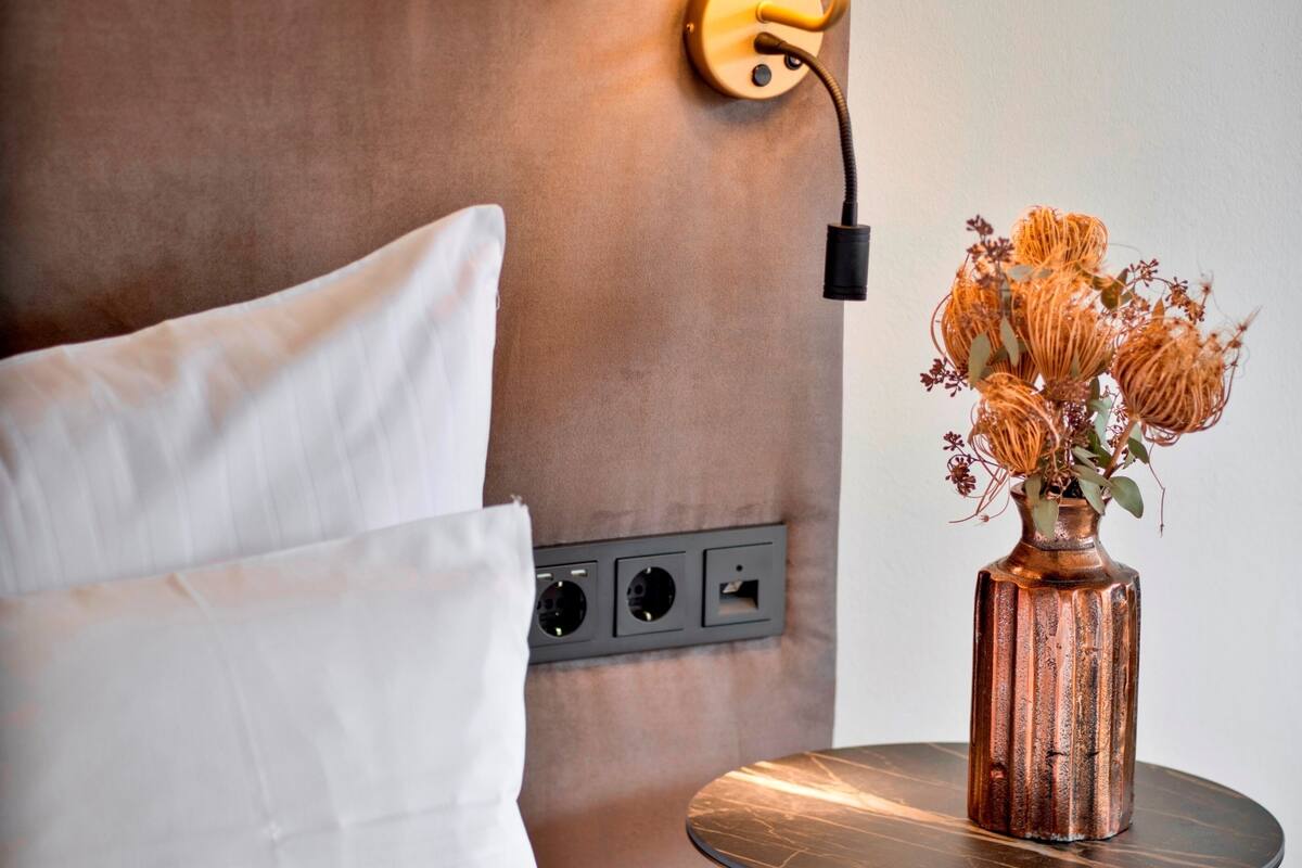 Une chambre d’hôtel avec une tête de lit, une lampe, une prise électrique et un vase contenant des fleurs séchées sur une table de chevet.