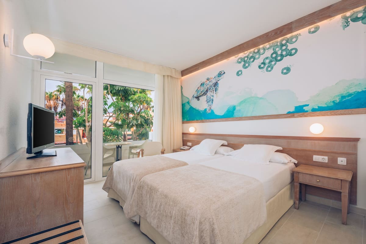 A hotel room with two beds, a TV, a desk, and a large window with a view of palm trees.